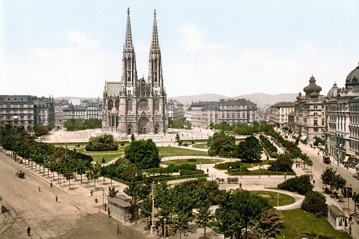 A colour print from about 1900 showing the twin-towered Votivkirche across an open square with trees, paths and tall city buildings.