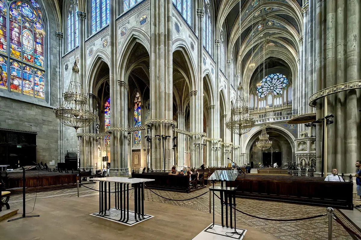 The nave of the Votivkirche in ordinary light, with tall pointed arches, grey stone piers, a blue and red stained-glass window at the left and the chancel in the distance.