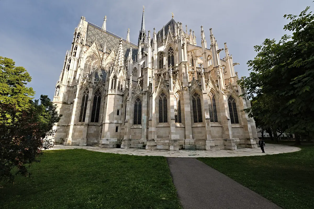 The east end of the Votivkirche seen from the surrounding park, with tall windows, buttresses and pinnacles in pale stone above a green lawn.